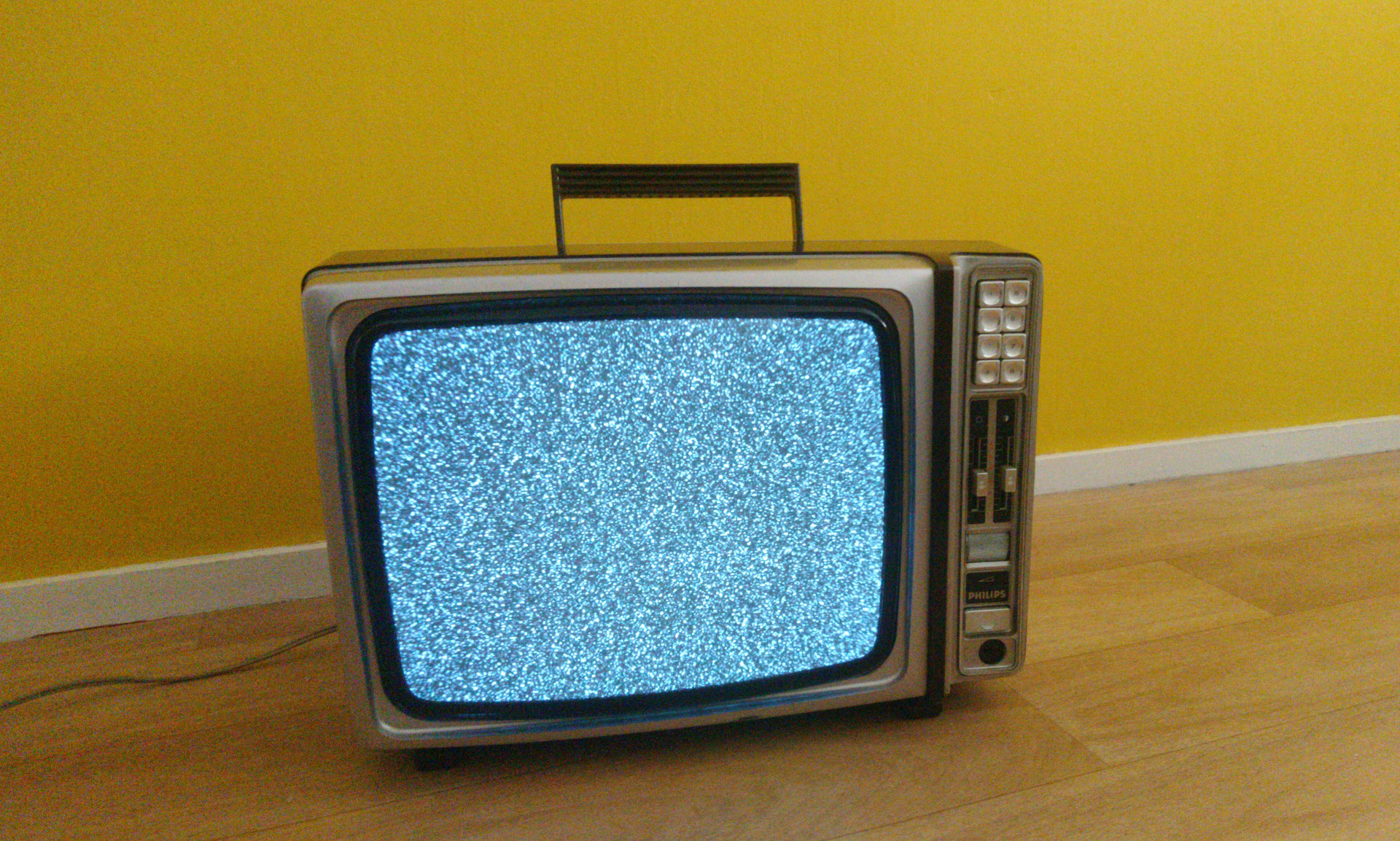 Télévision des années 70 grise et marron sur un faux plancher et devant un mur jaune. Munie d'un écran cathodique bombé aux rebords noirs affichant de la neige sur la gauche et des commandes sur la droite. Le panneau de contrôle comporte de haut en bas : 8 boutons poussoirs sur deux colonnes dont on distingue uniquement les numéros 1, 4, 5, 6, 7 et 8 ; 2 faders verticaux l'un à côté de l'autre pour régler la luminosité et le contraste ; Une molette texturée pour régler le volume ; L'inscription « Philips » ; Un bouton poussoir de mise sous tension ; Une prise casque jack.